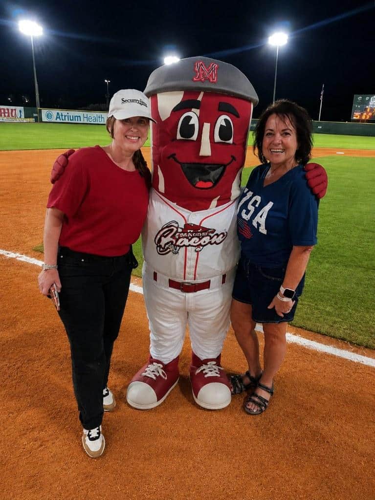 Two guests in Security First Asset Management caps pose with the Macon Bacon mascot on the infield under the stadium lights.